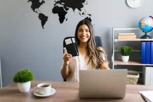 India e-Visa for New Zealand Travellers - Mann Travel Smiling woman sits at a desk in a modern office, holding a passport with boarding passes, laptop open in front, world map on the wall behind, globe and bookshelf to the side.