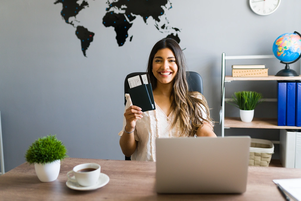 Smiling woman sits at a desk in a modern office, holding a passport with boarding passes, laptop open in front, world map on the wall behind, globe and bookshelf to the side.