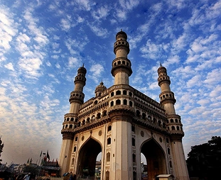 Charminar, a historic four-minaret gateway in Hyderabad, rising over a stone arch against a blue, partly cloudy sky.